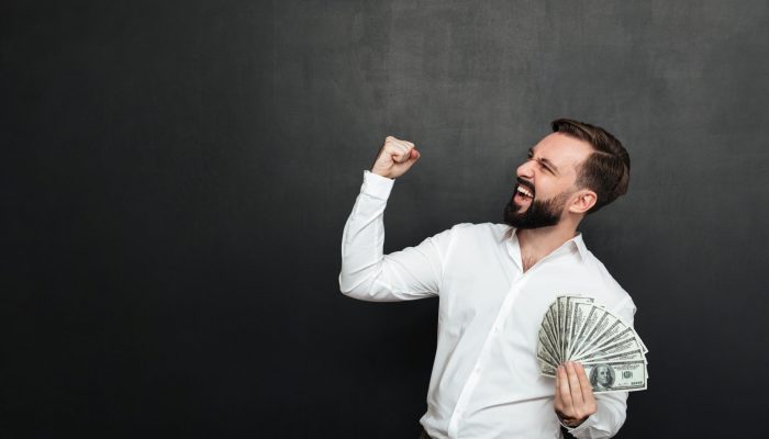 Portrait of successful guy in white shirt rejoicing like winner with fan of 100 dollar bills in hand clenching fist aside over dark gray background
